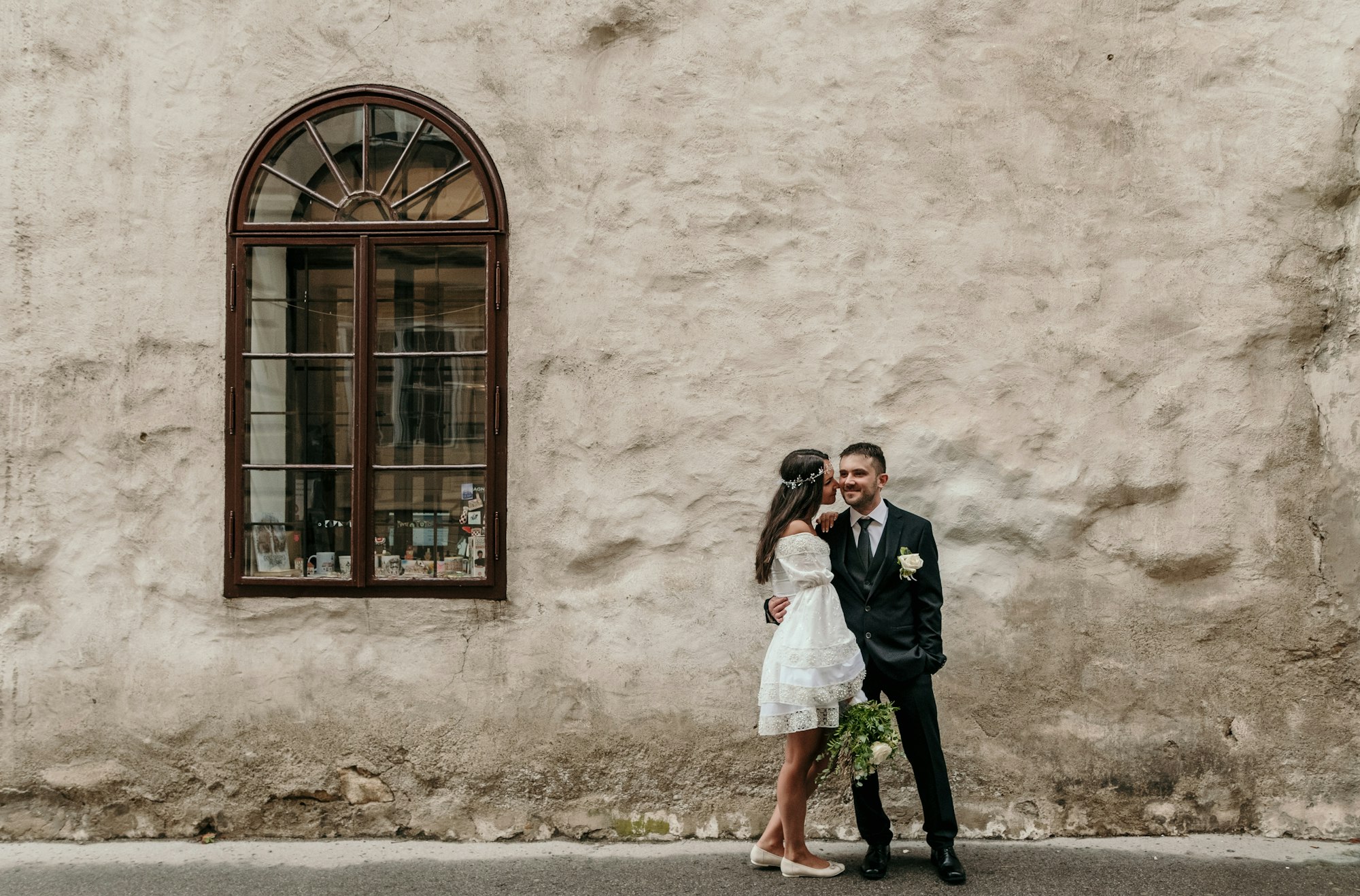 Minimalist boho style wedding photo of bride and groom in front of weathered wall in city.